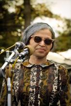 Photo of Rosa Parks with sunglasses on and speaking at a podium.
