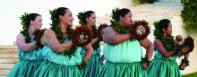 Women in blue dresses holding feathered gourds perform a traditional Hawaiian dance.