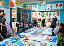 Students stand around a table with art work while a teacher holds up another piece of art work for them to see