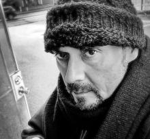 Black and white photo of man with a goatee wearing wool hat and a heavy coat standing in doorway. 