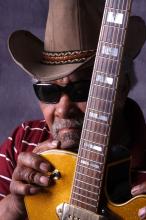 A man in sunglasses and a cowboy hat holds his guitar in a close-up photo