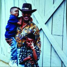 Man in black hat holding a boy in his right hand with another boy standing in front of him. 