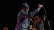 Black man in hat and shiny jacket singing into mic on stage. 