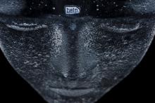 detail from a mask which is painted black and has a scrap with the word help embedded in the forehead of the mask