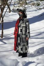 Woman standing in snow wearing hat, coat, scarf, and mask with Native American motifs.