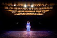 A woman in a blue dress stands on a stage facing a dark auditorium