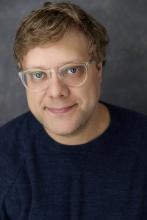 Headshot of Andrew Dolan, a white man with sandy blonde hair, glasses, and a blue eyes. He wears a grey shirt. 