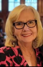 an older white woman with shoulder length blonde hair and glasses smiles gently at the camera