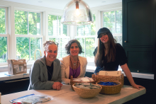 two white women sit around a kitchen table while another stands near the table