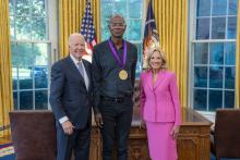 Tall bald Black man wearing glasses with a medal around his neck surrounded by older man wearing a blue suit and woman with blonde hair wearing a pink suit. 