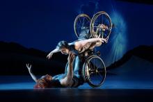 Laurel Lawson, a white woman with short cropped teal hair, is flying in the air with arms spread wide, wheels spinning, and supported by Alice Sheppard. Alice, a multiracial Black woman with coffee-colored hair, is lifting from the ground below. They are making eye contact and smiling. A burst of white light appears in a dark blue sky. 