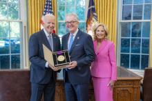 Man with gray hair and glasses holding a medal surrounded by older man wearing a blue suit and woman with blonde hair wearing a pink suit. 