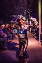 Man walking the runway as a futuristic cyborg, wearing glasses a futuristic silver costume. Audience members are gathered on both sides of the runway.