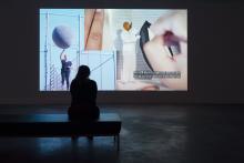 Woman sitting on a bench in a dark room, facing an art exhibit that depicts a woman holding a large ball and a painting of a black man in a white suit, with text in the exhibit that says: "I'm still here in spite of efforts to displace me and my community."