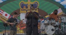 On a music stage: Black man (center) playing a trumpet, White man (left) playing the guitar, and Black man (right) playing drums.