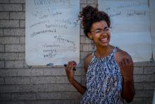 photo of Kendra Kreuger, who is a young Black woman with her hair pulled back and wearing glasses. She stands in front of large sheets of paper covered with text.
