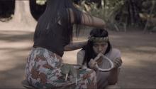 Film still of two women: Woman on left is tying a headpiece on to the woman on the right. Woman on the right is kneeling and playing an instrument.