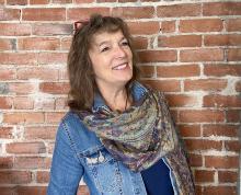 a middle-aged woman wearing a denim jacket and scarf, photographed against a brick wall