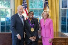 Black woman in dark blue suit wearing a medal around her neck surrounded by older man wearing a blue suit and woman with blonde hair wearing a pink suit. 