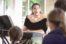 Woman wearing black dress and large gold necklace sitting at piano and singing in front of an audience. 