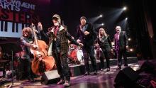 Three men and a woman smile and walk single-file onstage while musicians play behind them.