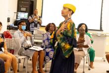 Black woman in yellow hat and flowery shirt stands in foreground with hand on her heart, group of people seated behind her also have hands over the heart, Black man at back plays a trumpet. 