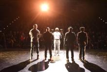 Five people (one standing center) stand on stage, facing a large audience.