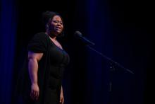 A young woman in a black dress speaks at a microphone