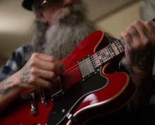 A man with a long grey beard and mustache sits behind a red guitar. His tattooed hands in the midst of playing a tune