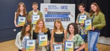 A group of teenagers smile and pose in front of a sign for the Musical Theater Songwriting Challenge. They each hold a binder with their name and the name of their winning song.