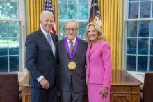 Man with glasses and gray hair and beard in gray suit wearing a medal around his neck surrounded by older man wearing a blue suit and woman with blonde hair wearing a pink suit. 