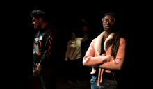 On stage: Male actor (left) is standing onstage with a black hoodie, female actor (middle) is wearing a white shirt and holding a red cup, and female actress (right) is wearing a pink jacket and heart-shaped glasses and crossing her arms