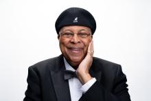 Latino man wearing tuxedo and bow-tie, with beret, left hand holding his face, smiling.