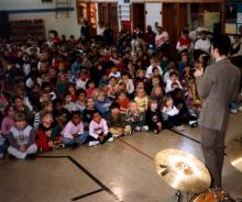 A large group of young children sitting cross-legged on the floor listening to a man play a saxophone