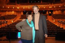 a woman in a brown shirt and a blue scarf stands on a stage with a man with a beard wearing a brown suit.