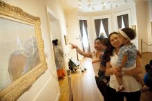 two women holding their toddlers and looking at a landscape painting on the wall of a museum