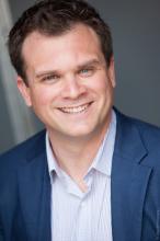 a white man with dark hair and blue eyes in a blue striped collared shirt and blue sportcoat