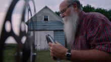 an older man with a long white beard stands outside a barn loading a reel onto an old fashioned film projector
