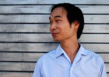 headshot of smiling Asian man in half-profile against background of weathered wood siding