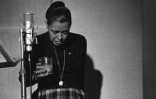 Billie Holiday in black turtleneck stands behind a microphone with her head down and a drink in her hand.