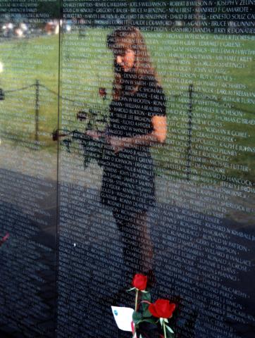 Reflection of a woman in a dress standing in front of a black reflective monument with names engraved on it, a flower at her feet. 