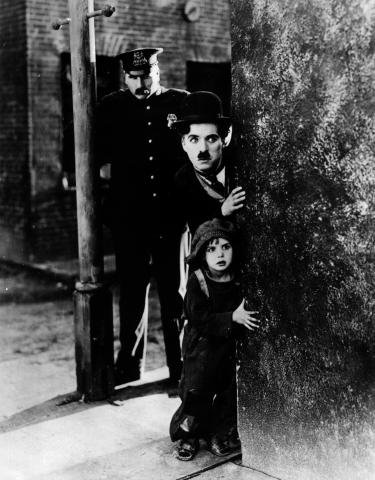 black and white still of man in a hat with a small black moustache with a little boy below him, both looking around a corner, with a police officer behind them. 