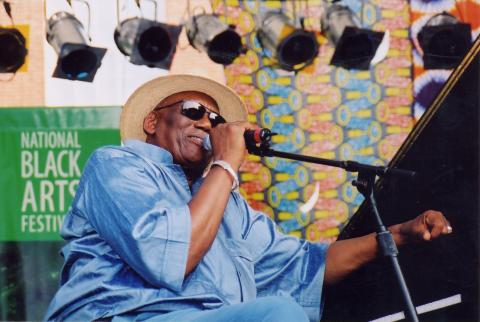 Black man wearing straw hat and sunglasses and blue shirt at piano speaking into a mic on an outdoors stage. 
