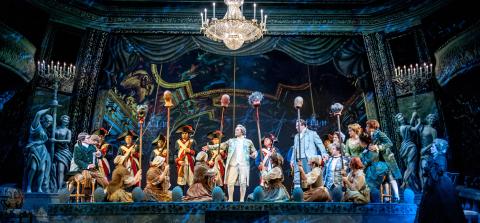 A stage production with a large cast dressed in 19th century clothing with man in the middle singing. 