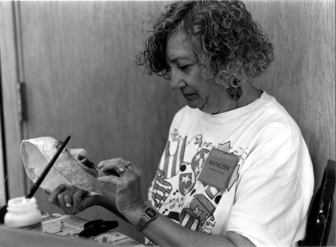 A white woman with long curly hair wearing a white tee-shirt is working on an art piece. 