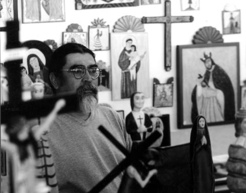 Man with glasses, dark hair, and gray goatee sitting in a studio with religious icons. 