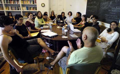 A large group of men and women of all ages sitting around a table listening to an older man in white shirt speak. 