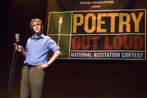 Male student in blue shirt standing in front of mic on stage with a large sign behind him. 