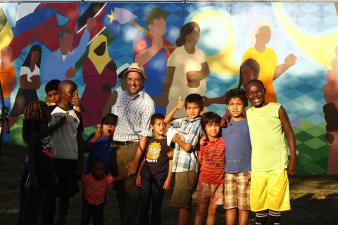 A group of children surrounding a man in a white shirt and hat in front of a mural they just painted. 