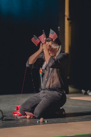 Man on stage kneeling holding three small US flags in front of his face.